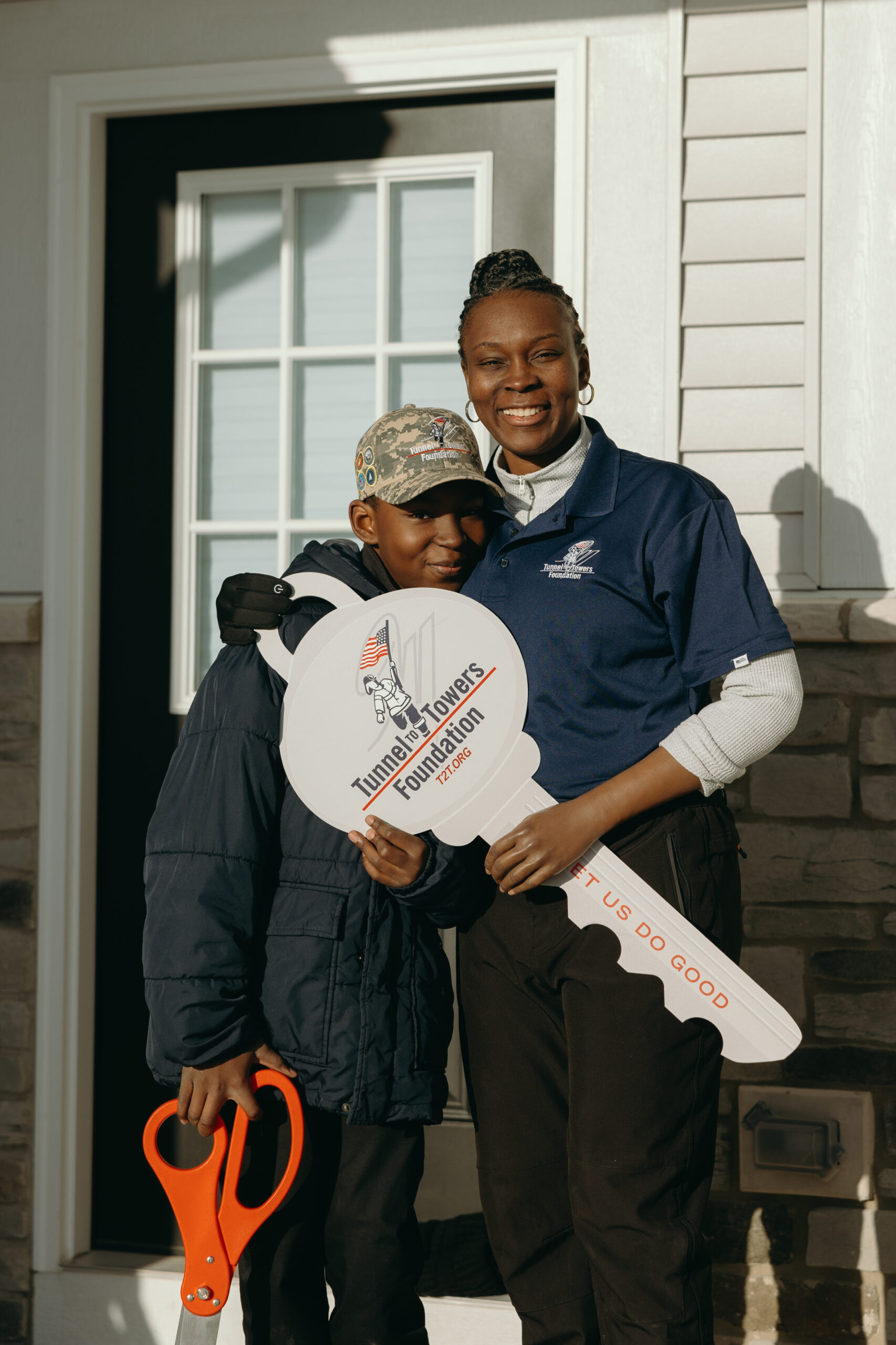 A woman holds a giant Tunnel to Towers key and a boy holds a giant pair of scissors in front of a house.