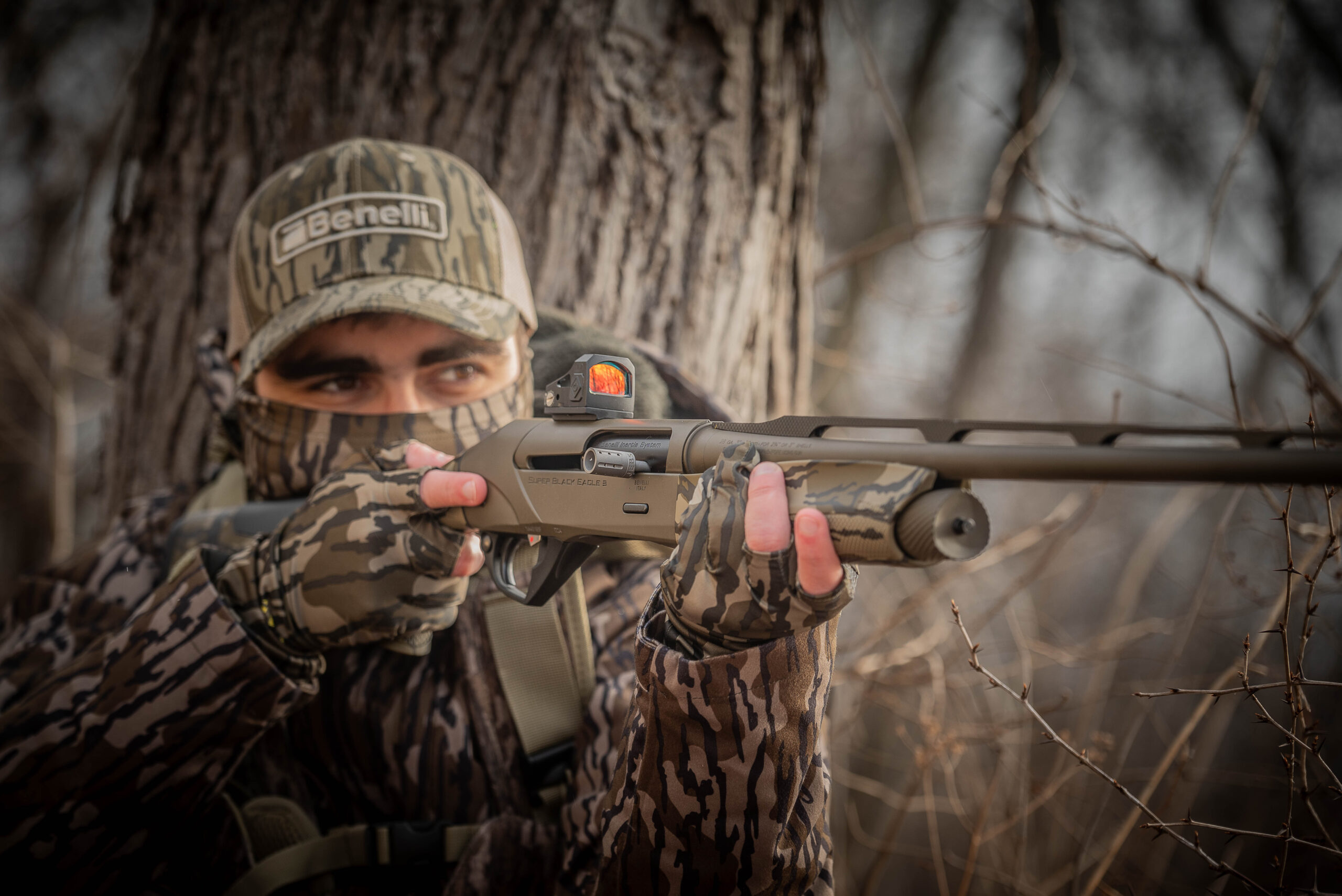A hunter wearing a Benelli cap and Bottomland camo aims a shotgun.