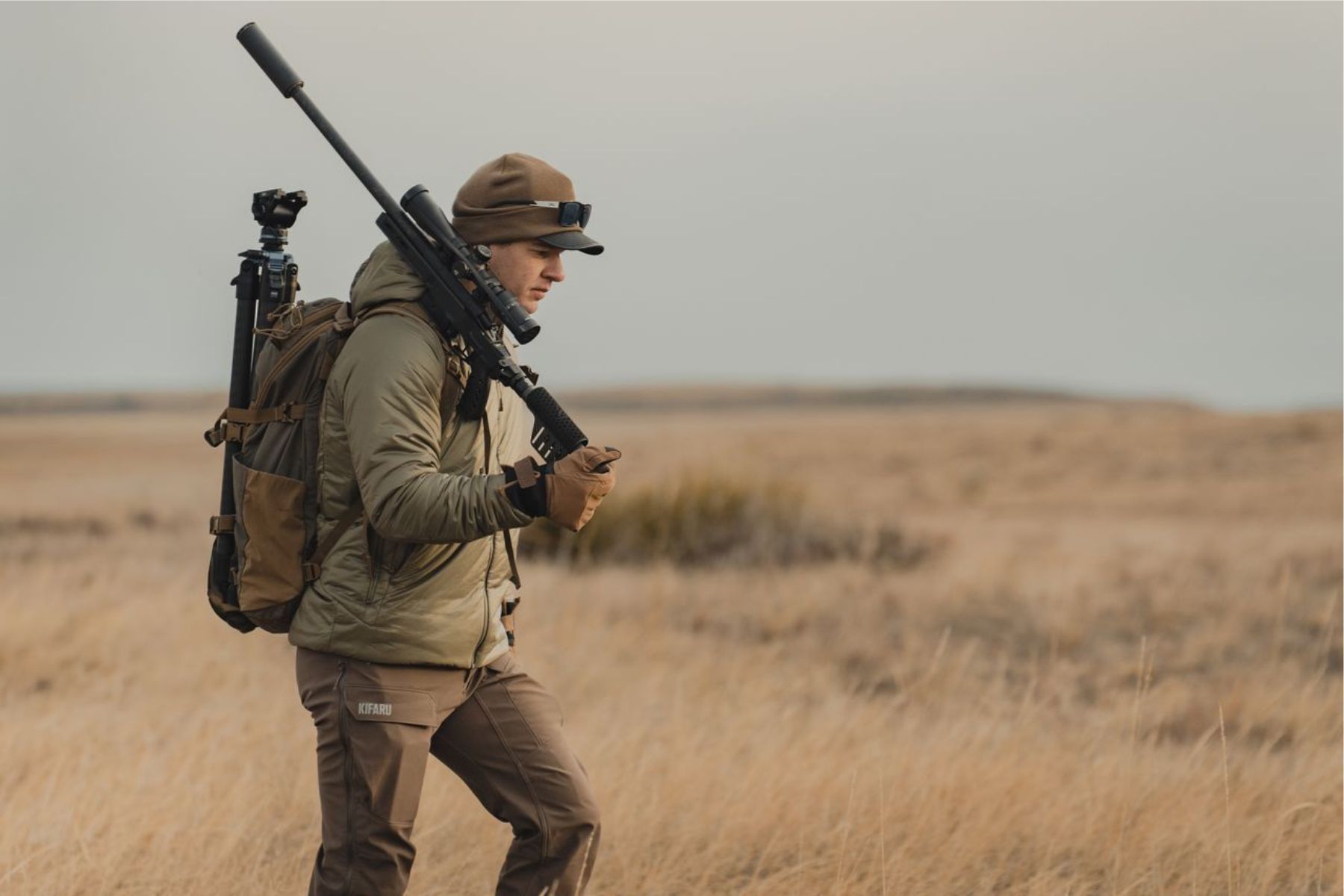 A hunter carrying a rifle across open grassland.