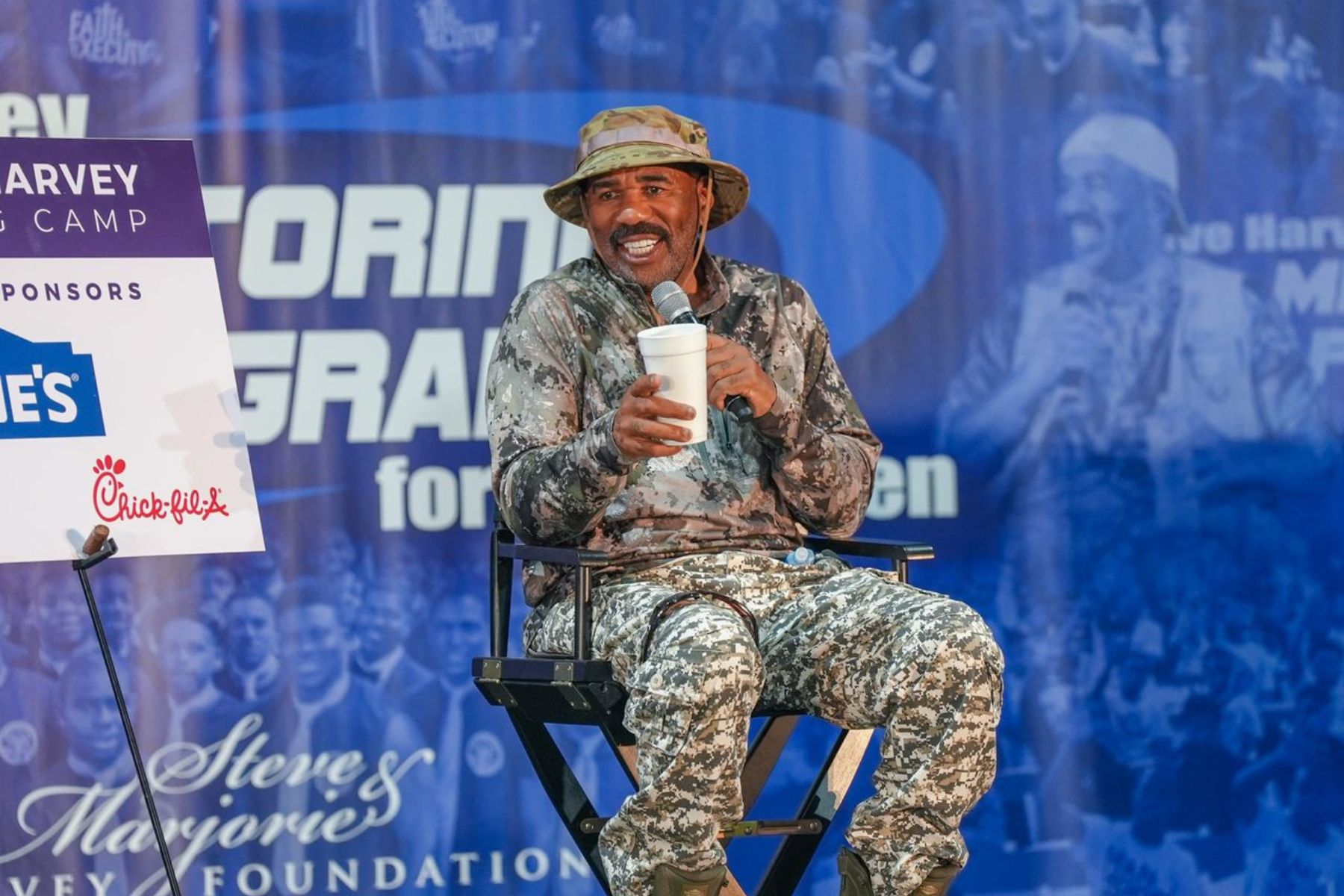 Steve Harvey sits in a directors chair and holds a microphone.