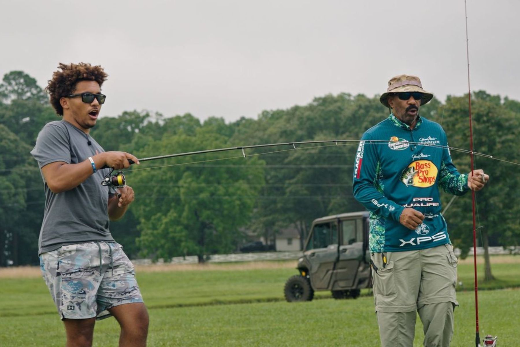 Steve Harvey stands next to a young man reeling in a fish.