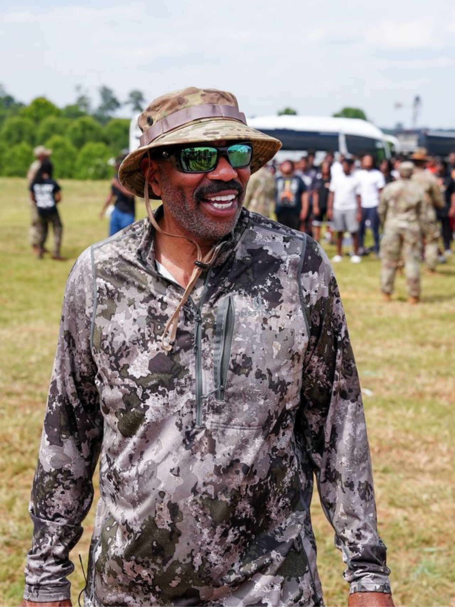 Steve Harvey smiles wearing a camo shirt and sunglasses.