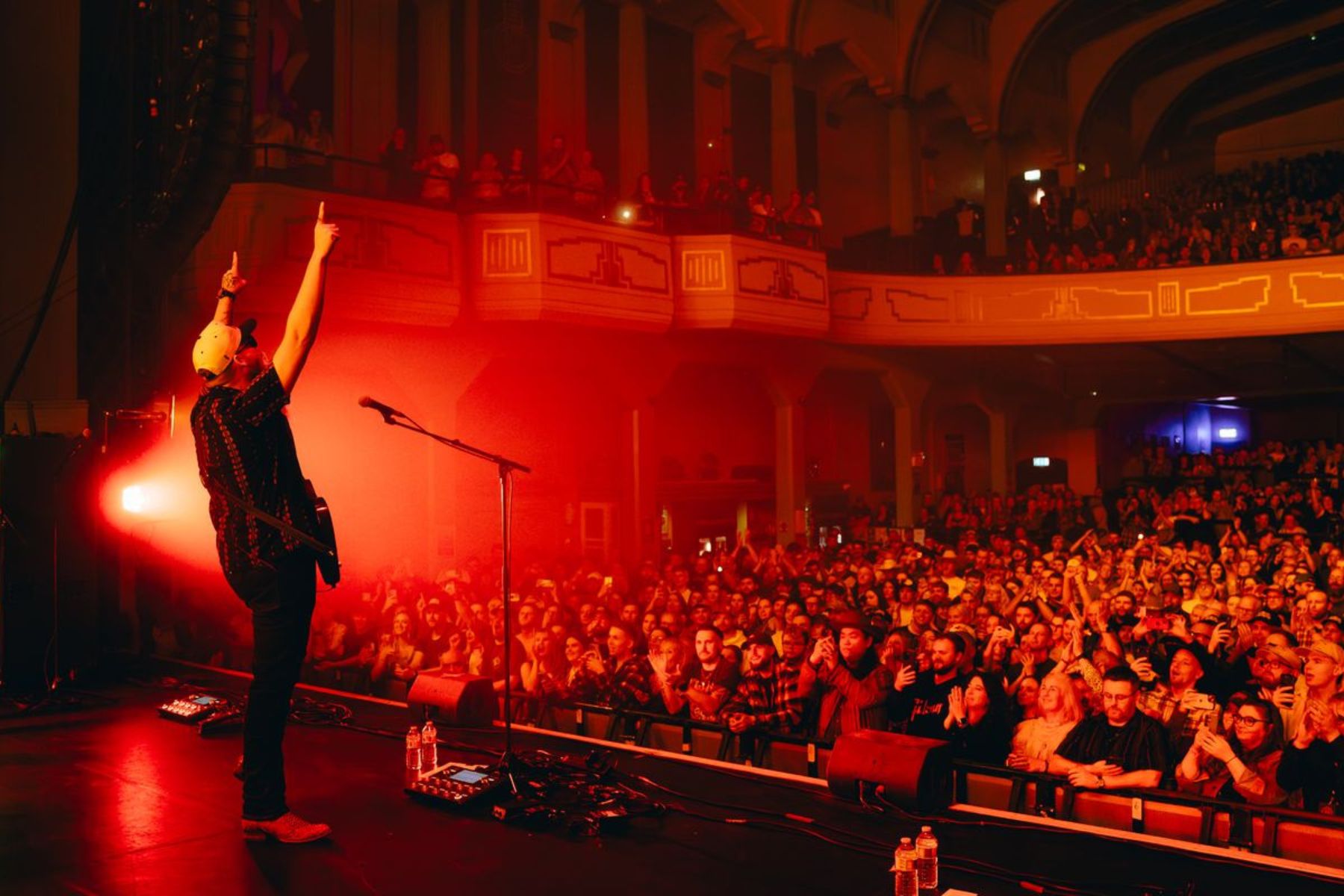 A man with a guitar points fingers to the ceiling in a crowded auditorium.