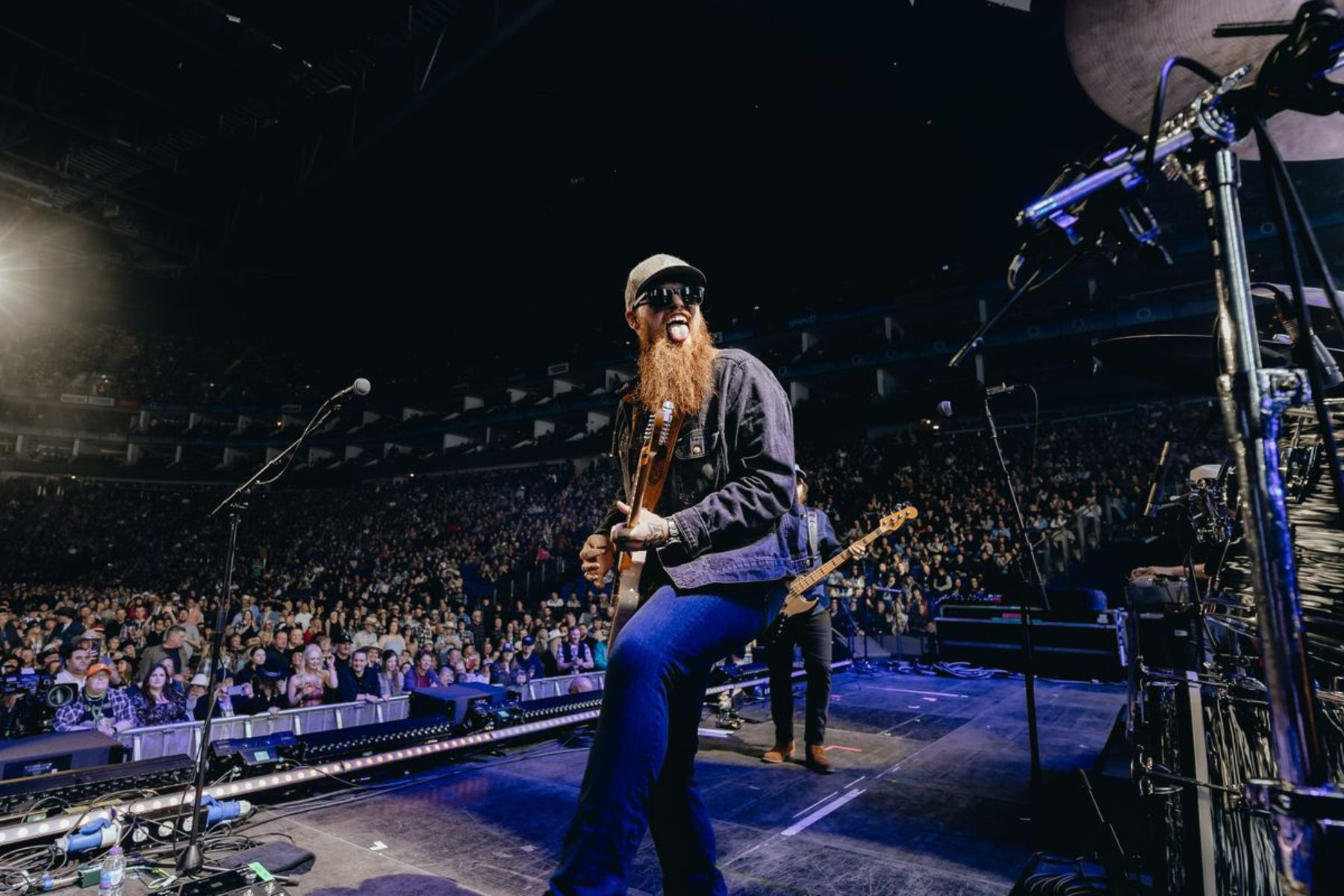 A bearded man sticks out his tongue and plays a guitar in front of a crowded auditorium,