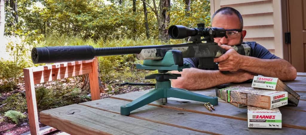 a man shooting a suppressed hunting rifle from a rest with boxes of Barnes Sierra Harvest Collection ammo in foreground