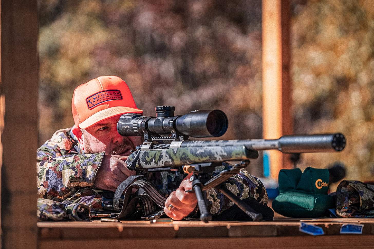a man in a FORLOH hat shooting a suppressed bolt action rifle with a high power scope from a tripod on a shooting bench