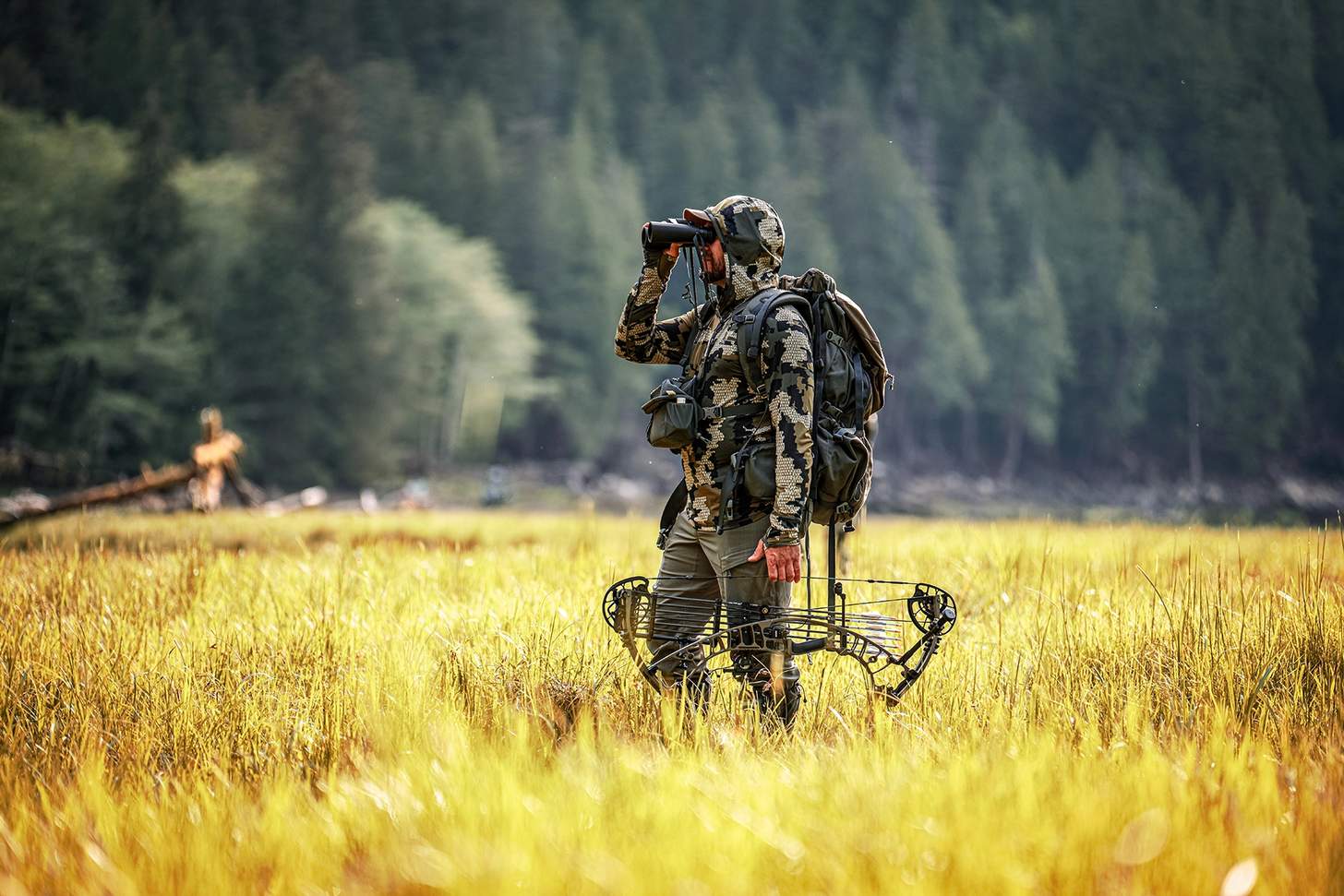 a hunter looking through binoculars holding a bow