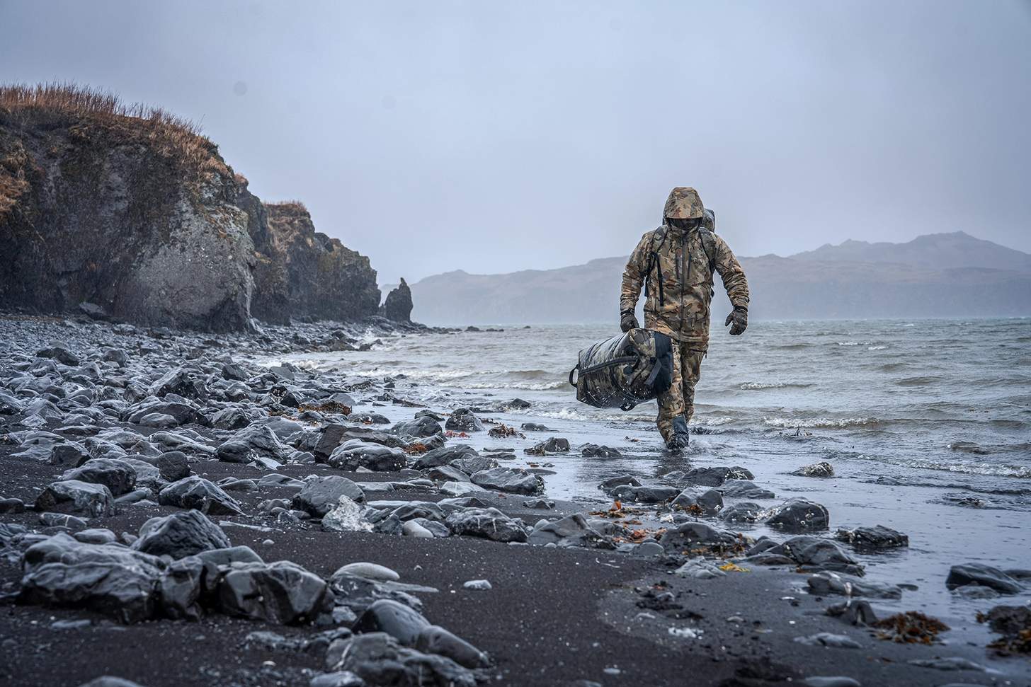 a hunter in kuiu gear walking along the shore with a gear bag