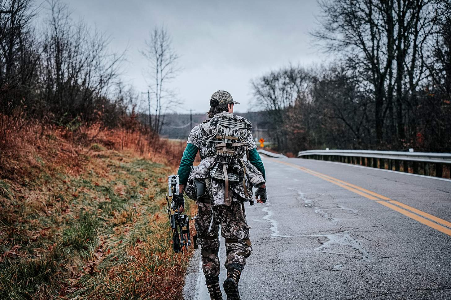 a bowhunter hiking on a road with his hunting saddle on his back
