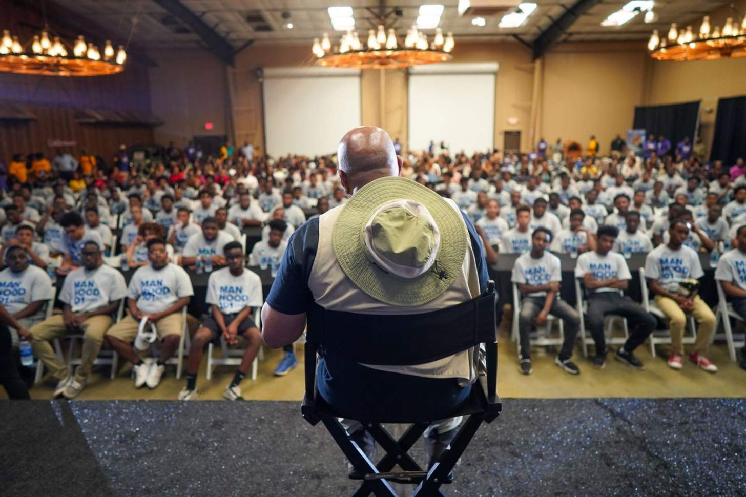 A bald man sits in front of an an audience made up of young men.