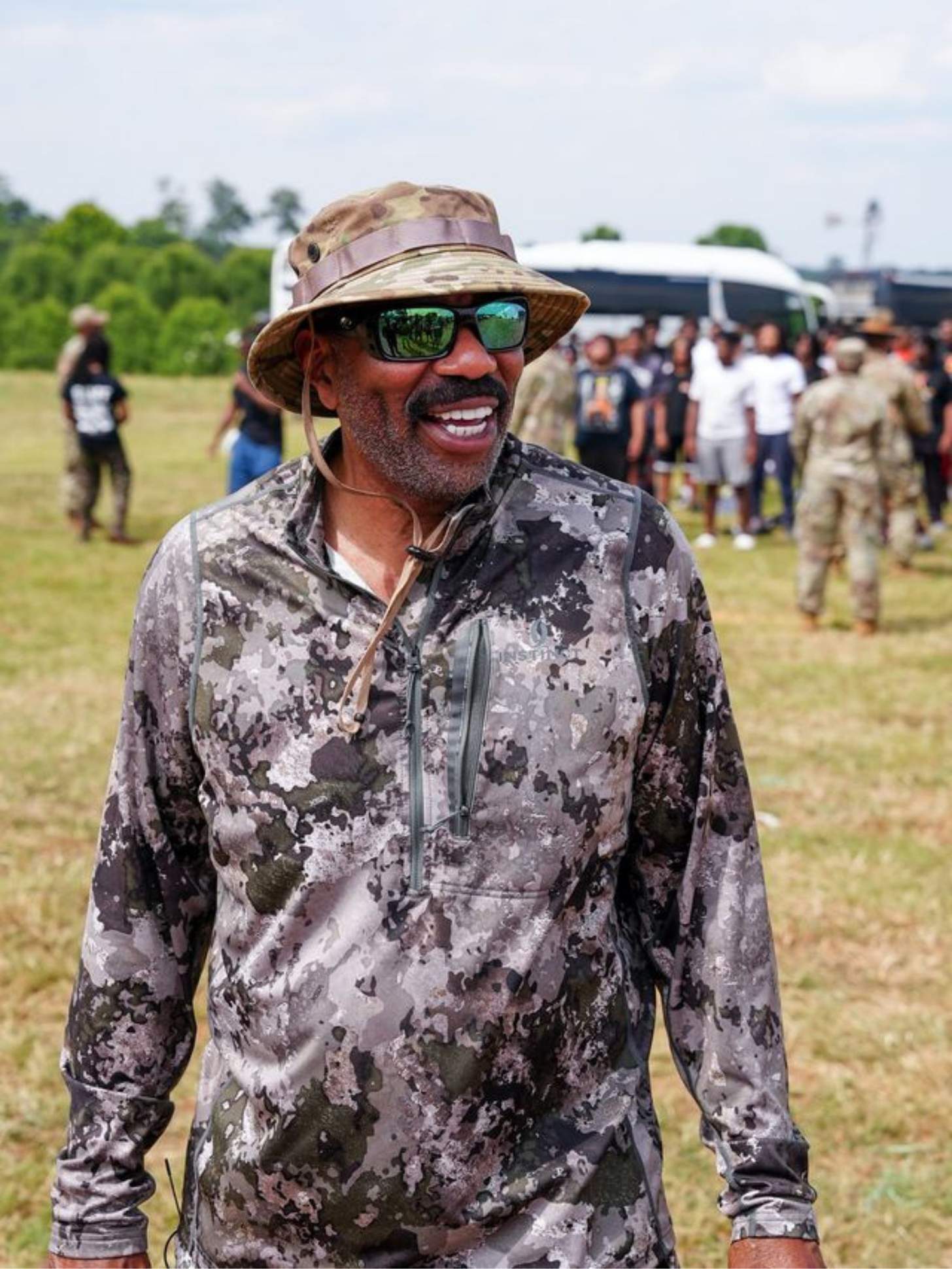 Steve Harvey smiles wearing a camo shirt and sunglasses.