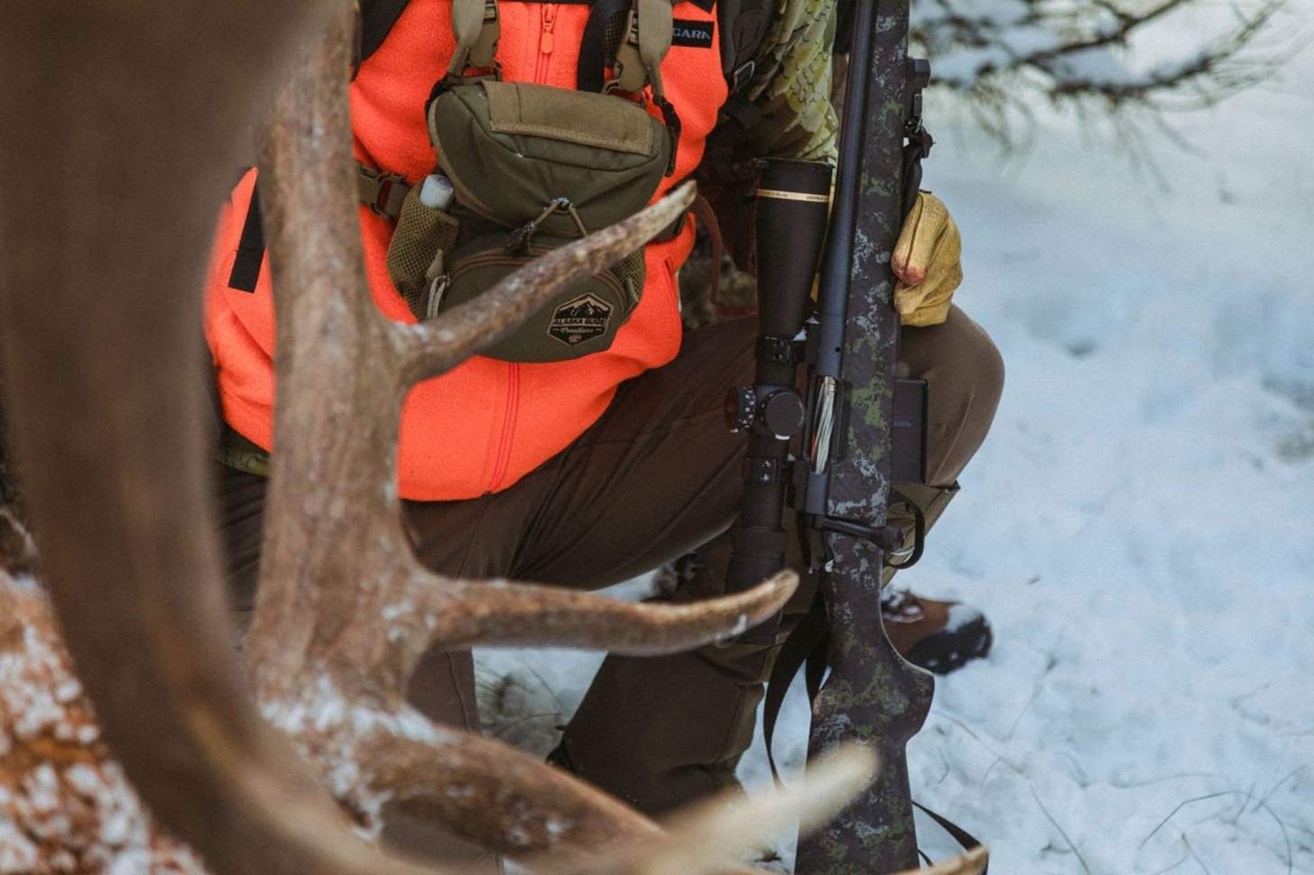 A hunter holds a rifle while standing behind deer antlers.
