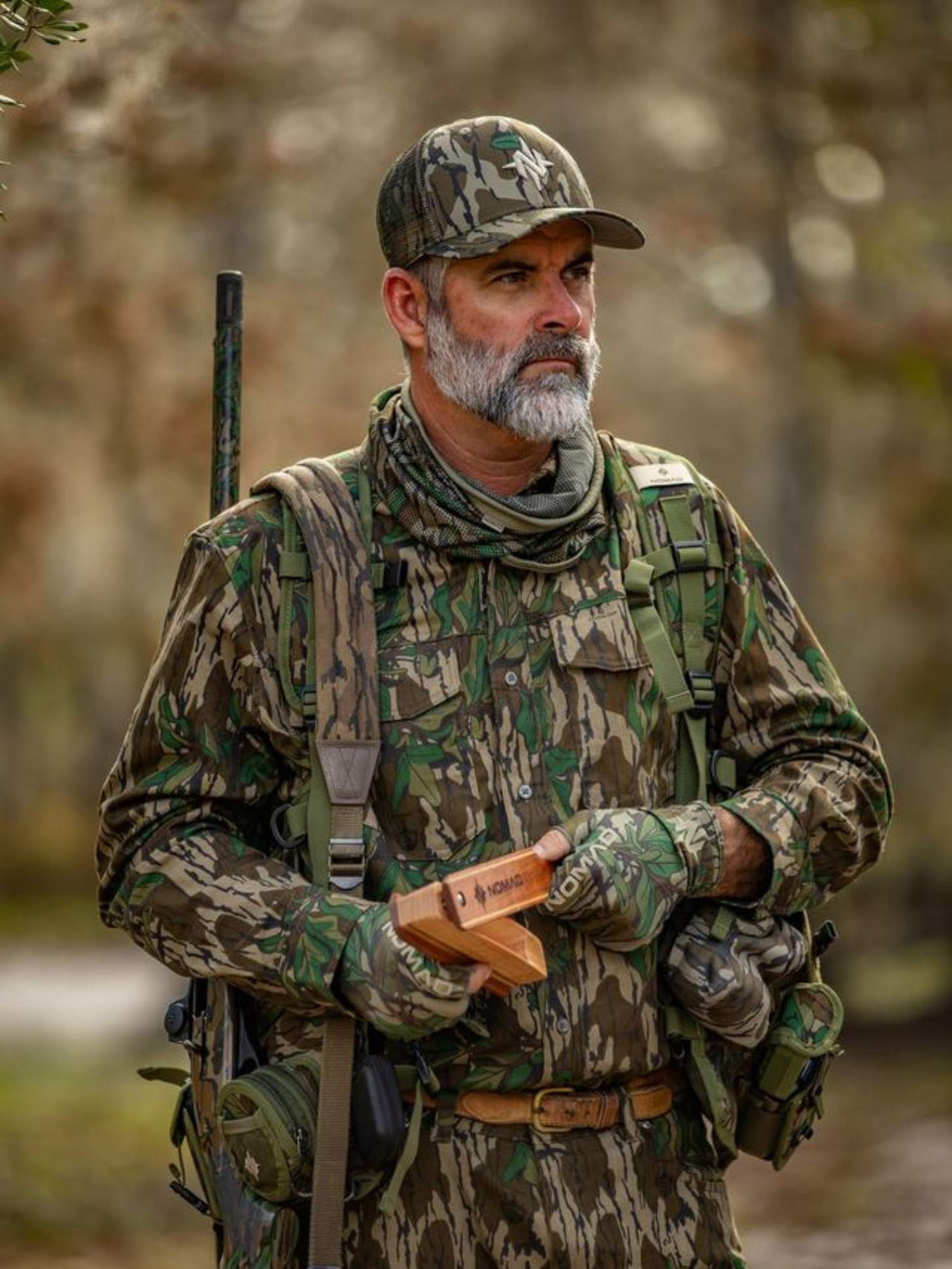 A man dressed in Mossy Oak Bottomland camo holds a shotgun and a box call.