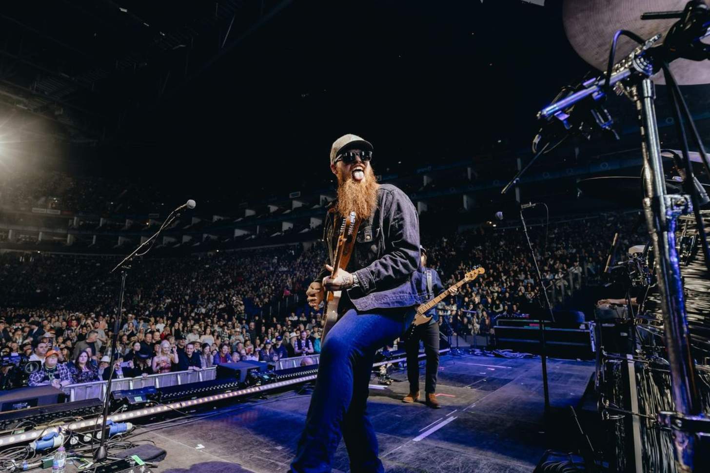 A bearded man sticks out his tongue and plays a guitar in front of a crowded auditorium,