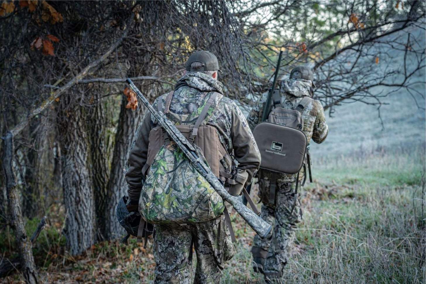 Two hunters dressed in camo and carrying shotguns walk along a wood line.