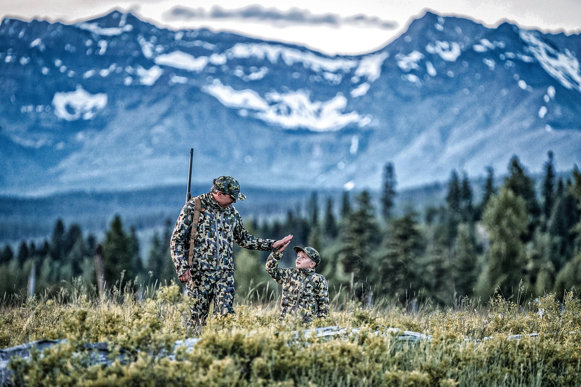 a father and son hunting in camo high-fiving in sprawling mountainous wilderness