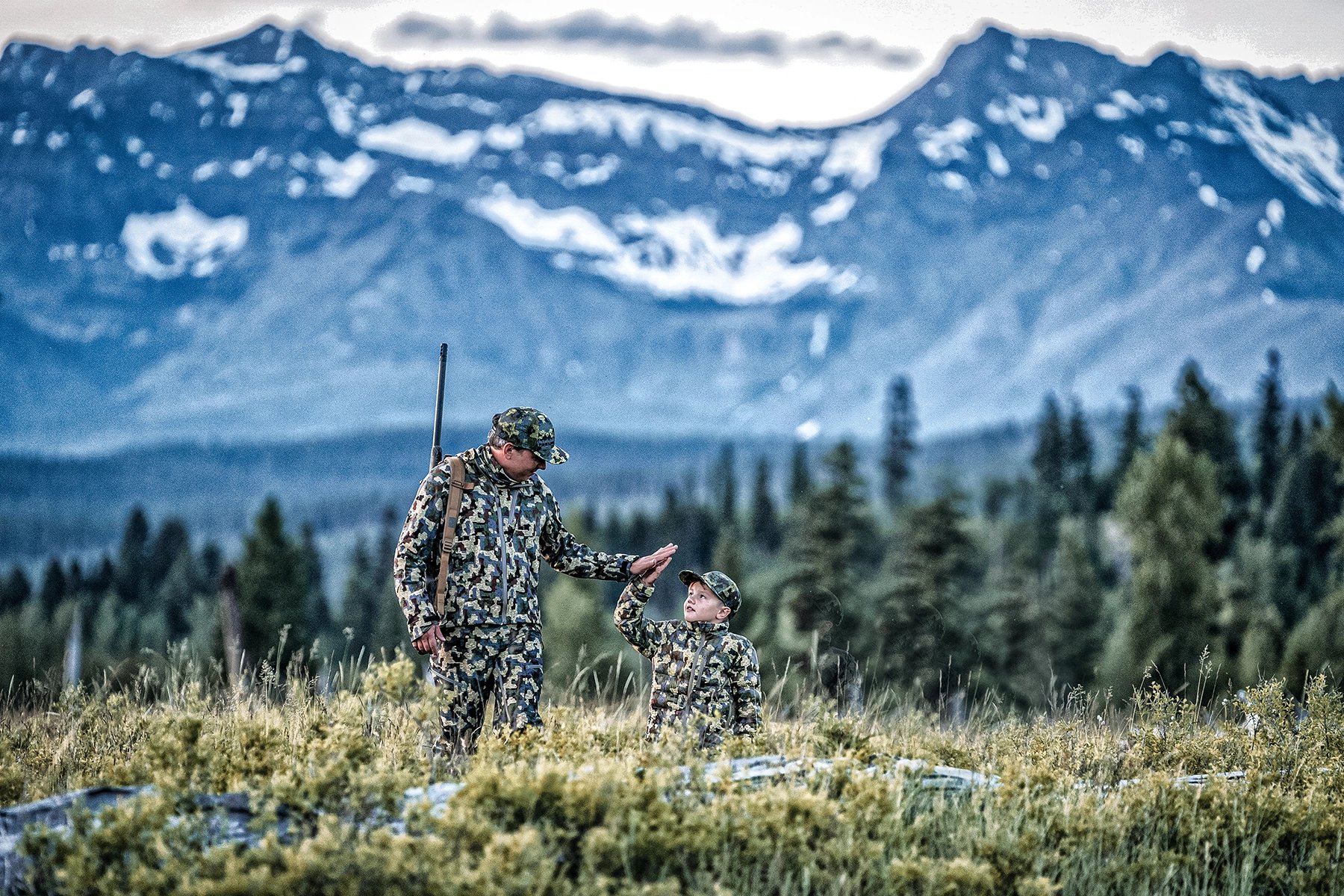a father and son hunting in camo high-fiving in sprawling mountainous wilderness