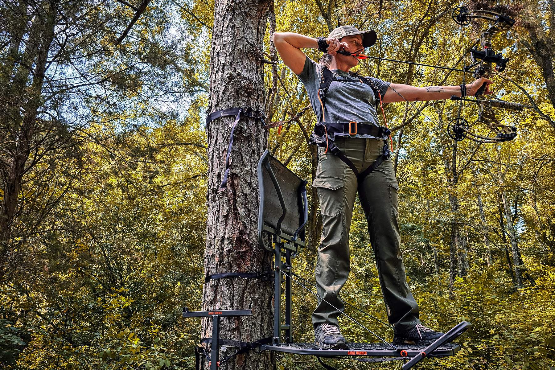 a female bowhunter taking practice shots with a compound bow from a treestand in summer