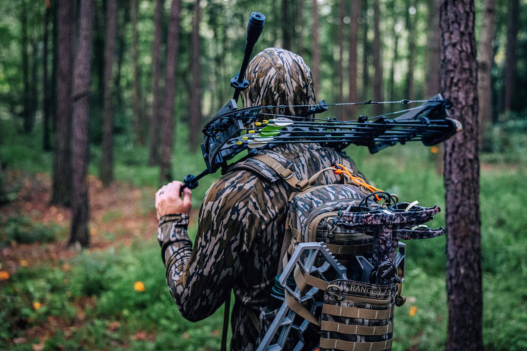 Born Primitive Apparel in Mossy Oak Bottomland bowhunting carrying compound bow and climbing stand